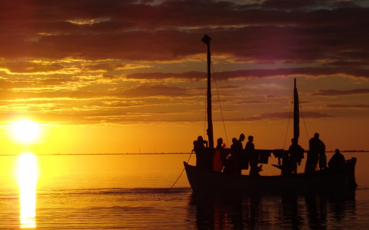 the silhouette of a sailboat holding a group of people is illuminated as the sun sets behind them. The sky appears in vibrant orange.